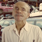 Man with a salt-and-pepper mustache and thinning hair, standing in front of an auto yard